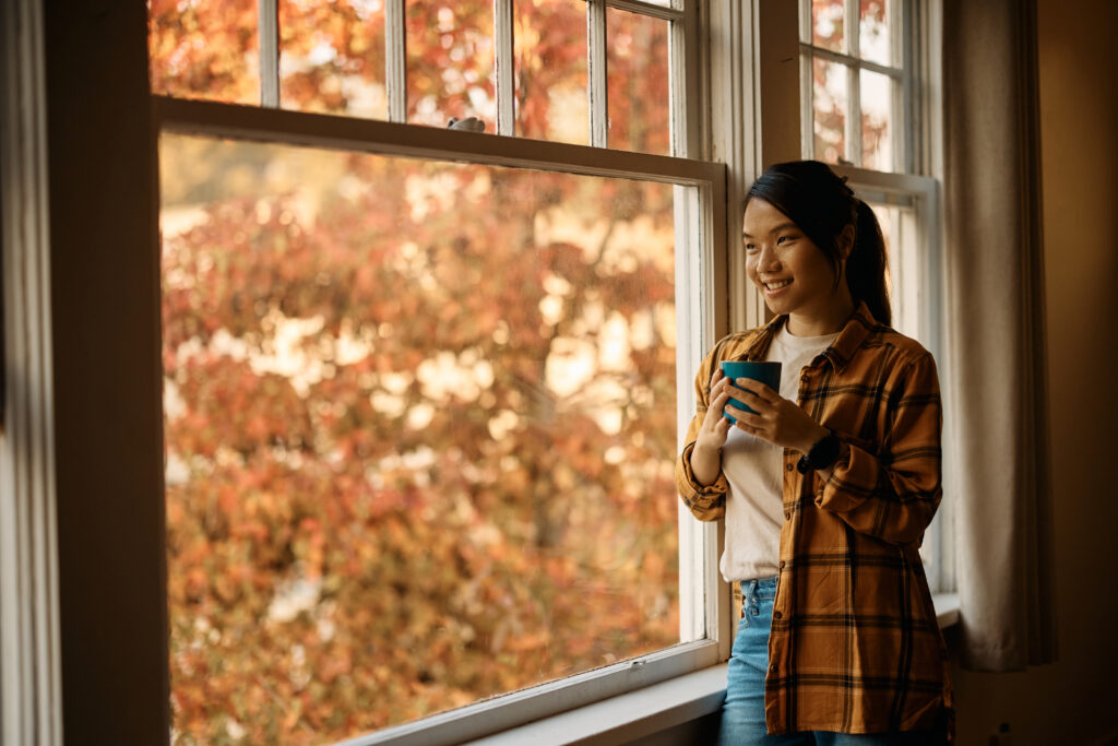 Smiling Asian woman having cup of coffee while looking through the window at home